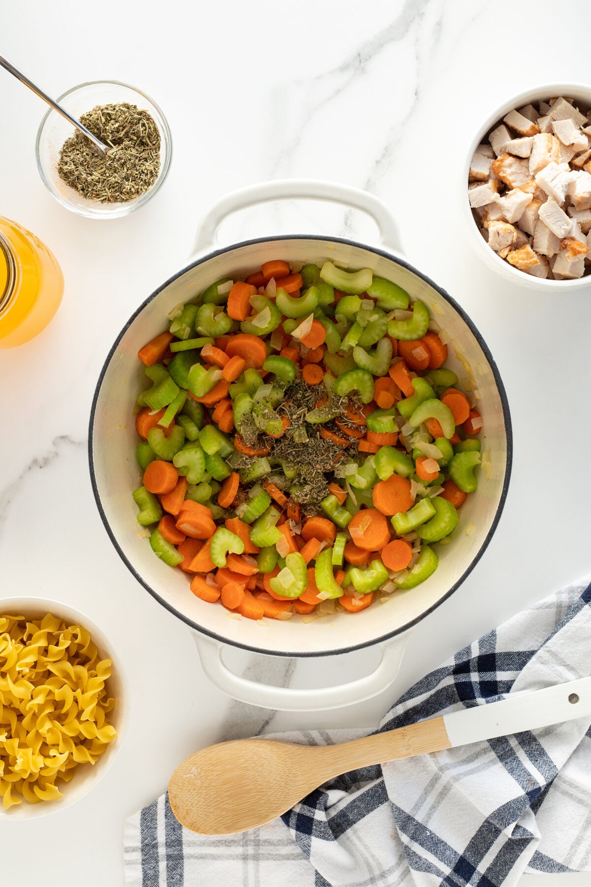 A white pot filled with chopped carrots and celery, topped with dried herbs, sits on a white counter. Surrounding it are a bowl of pasta, a bowl of cooked chicken cubes, a jar of broth, and a wooden spoon on a striped towel.