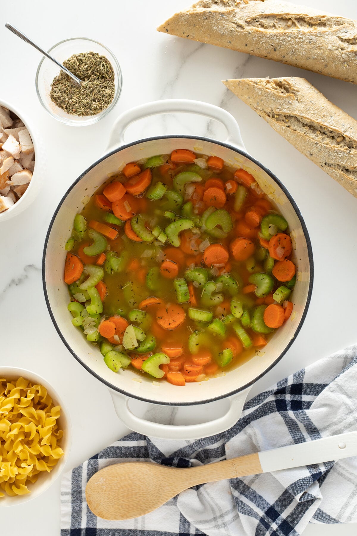 A pot of vegetable soup with sliced carrots and celery is on a white countertop, surrounded by a wooden spoon, a blue-and-white towel, a bowl of uncooked egg noodles, seasonings, chopped chicken, and two baguettes.