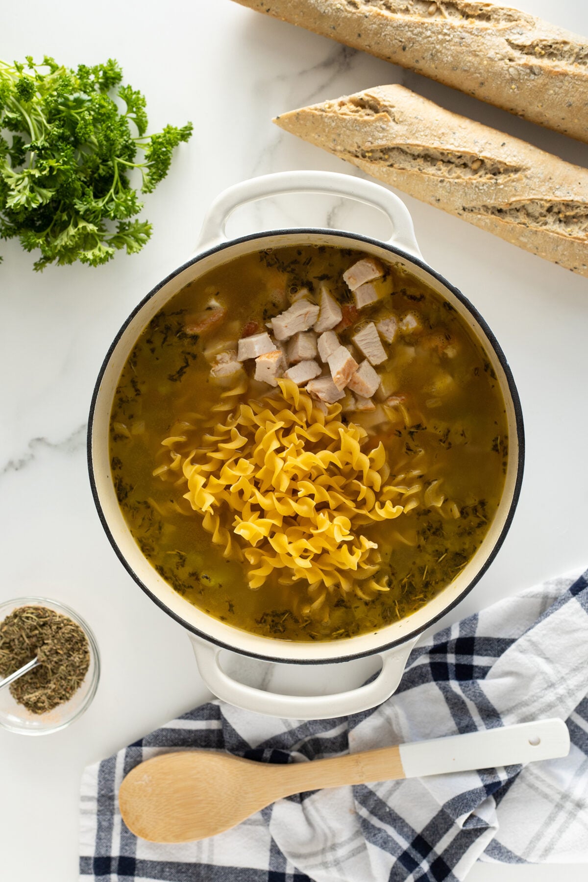 A pot of soup with uncooked ribbon noodles and diced chicken sits on a marble counter. Nearby are two baguettes, fresh parsley, a jar of seasoning, and a wooden spoon on a checkered towel.