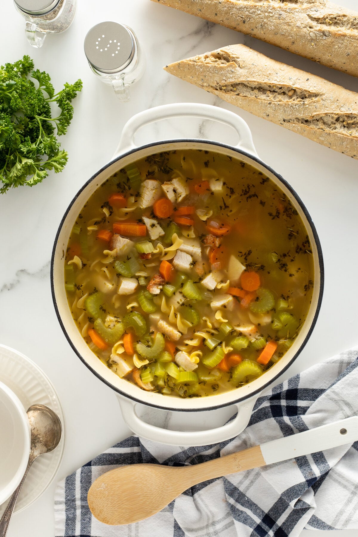 A pot of chicken noodle soup with carrots, celery, and herbs sits on a white surface next to a baguette, parsley, salt and pepper shakers, a wooden spoon, and a striped kitchen towel.