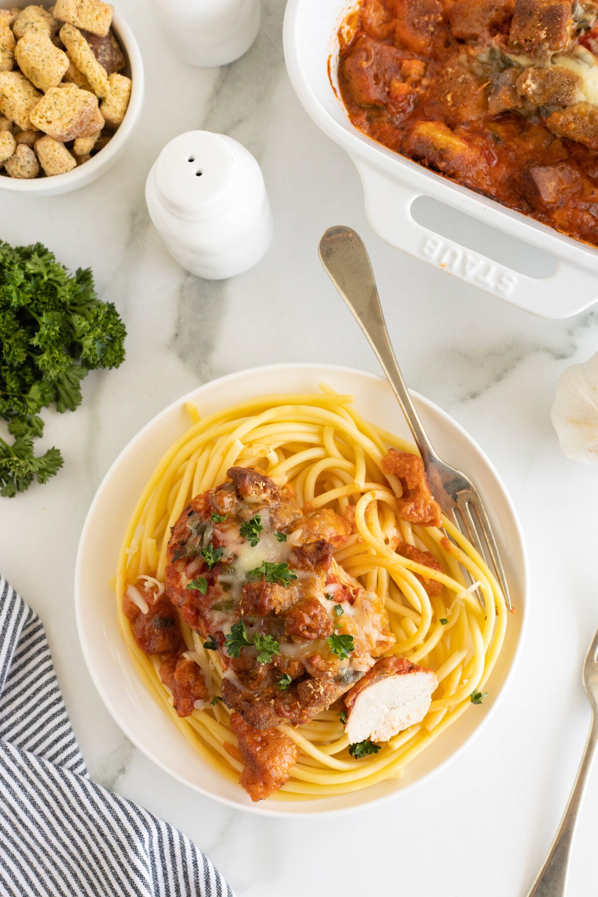 A plate of spaghetti topped with sliced chicken, tomato sauce, and herbs sits on a marble surface beside a fork, parsley, a dish of croutons, salt and pepper shakers, and a striped napkin.