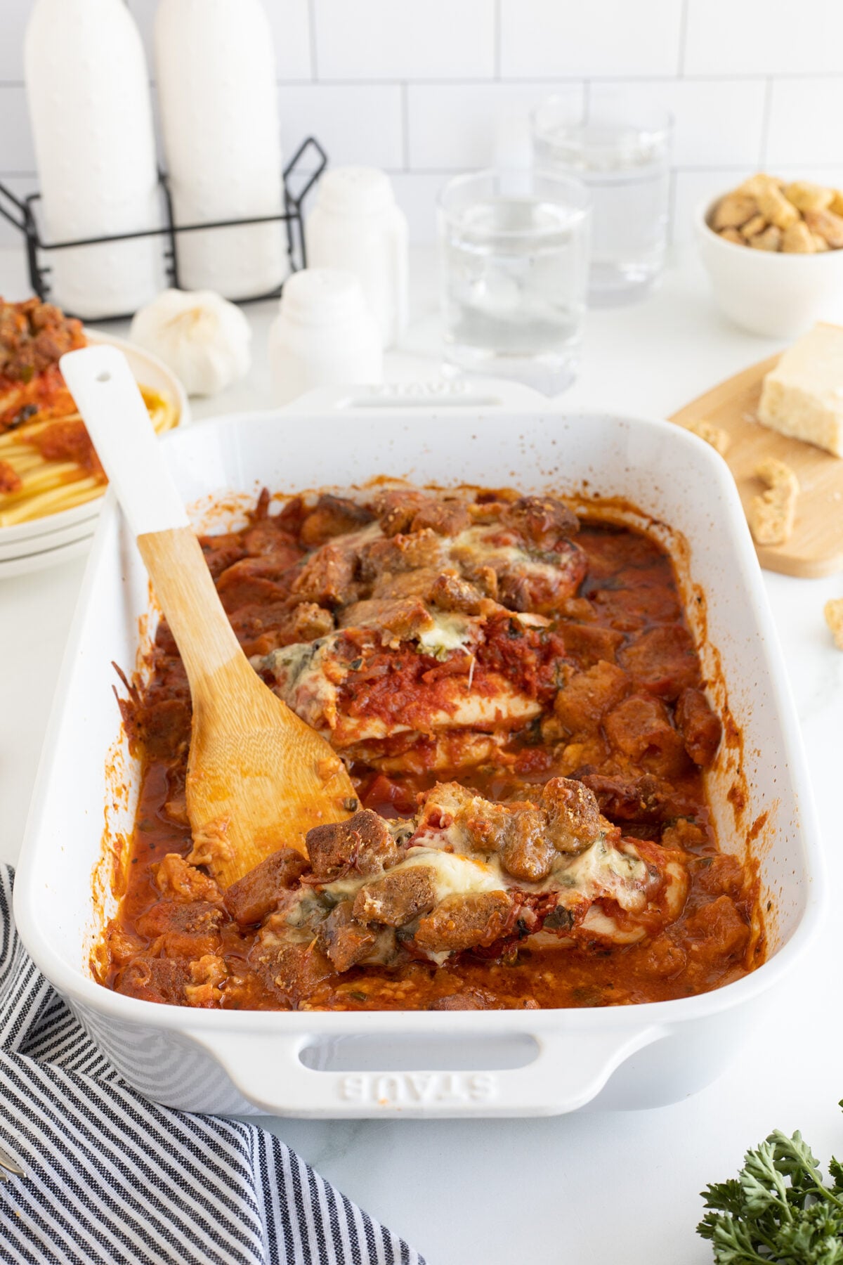 A white baking dish filled with baked chicken parmesan topped with melted cheese and tomato sauce. A wooden spoon rests inside, and the dish is set on a white table near water glasses, cheese, croutons, and seasonings.