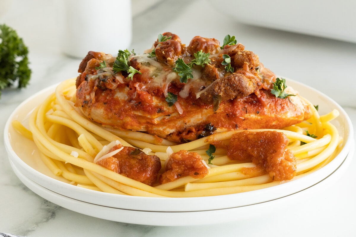 A plate of pasta topped with a piece of baked chicken, marinara sauce, chunks of tomato, melted cheese, and fresh parsley, served on a white dish.