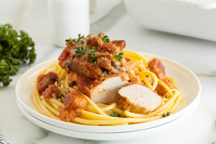A plate of pasta topped with sliced breaded chicken breast, tomato sauce, melted cheese, and garnished with fresh parsley. The dish is served on a white plate with herbs in the background.