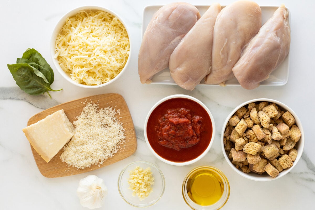 A top view of ingredients for chicken parmesan: raw chicken breasts, shredded cheese, basil leaves, grated parmesan, tomato sauce, croutons, minced garlic, olive oil, and a garlic bulb on a white surface.