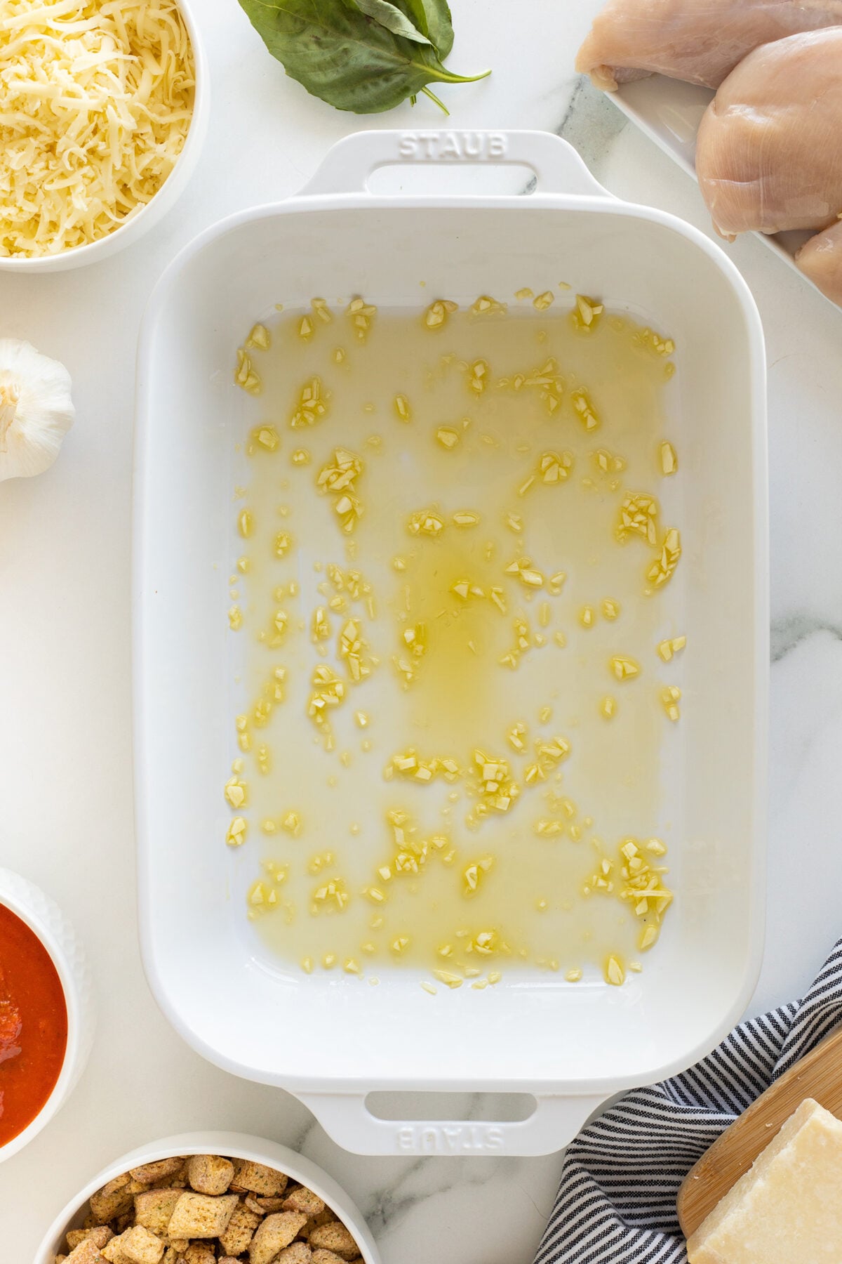 A white baking dish with minced garlic and olive oil spread on the bottom, surrounded by bowls of shredded cheese, croutons, marinara sauce, a garlic bulb, basil leaves, a wedge of parmesan, and raw chicken breasts.