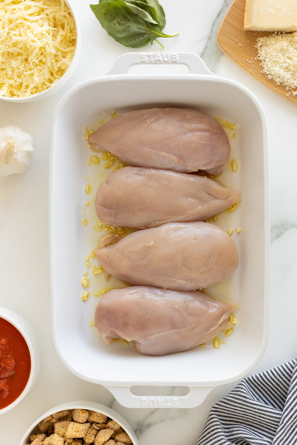 Four raw chicken breasts placed in a white baking dish with chopped garlic. Surrounding the dish are bowls of shredded cheese, tomato sauce, croutons, fresh basil, garlic, and a block of Parmesan cheese.