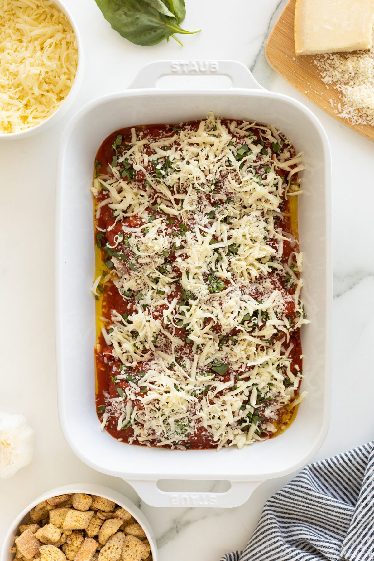 A white baking dish filled with a layered casserole topped with shredded cheese, herbs, and tomato sauce. Surrounding the dish are bowls of shredded cheese, croutons, garlic, fresh basil, and a cheese block on a board.