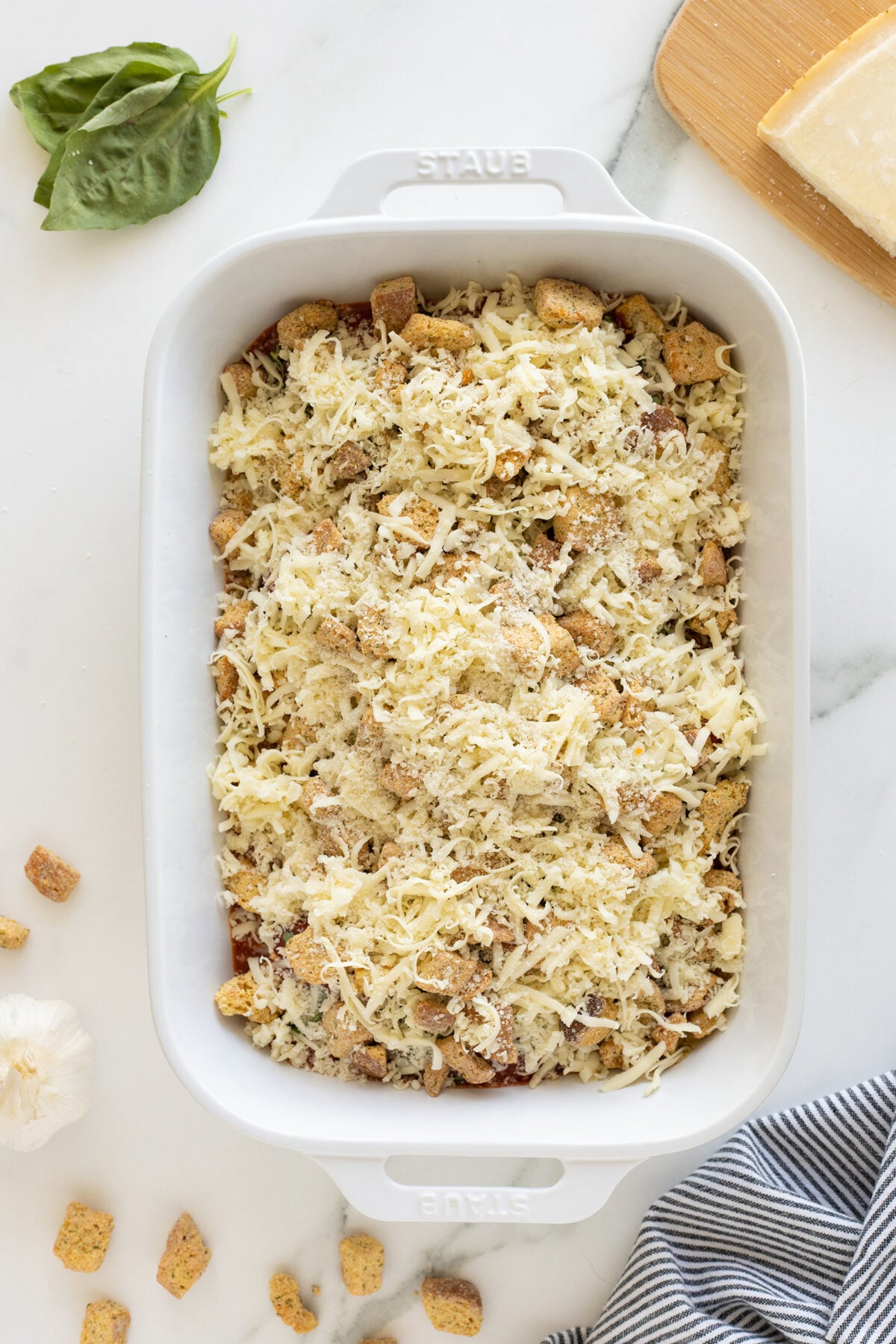 A white baking dish filled with an unbaked casserole topped with shredded cheese and croutons sits on a marble surface. Nearby are fresh basil leaves, a wedge of cheese, croutons, garlic, and a striped cloth.