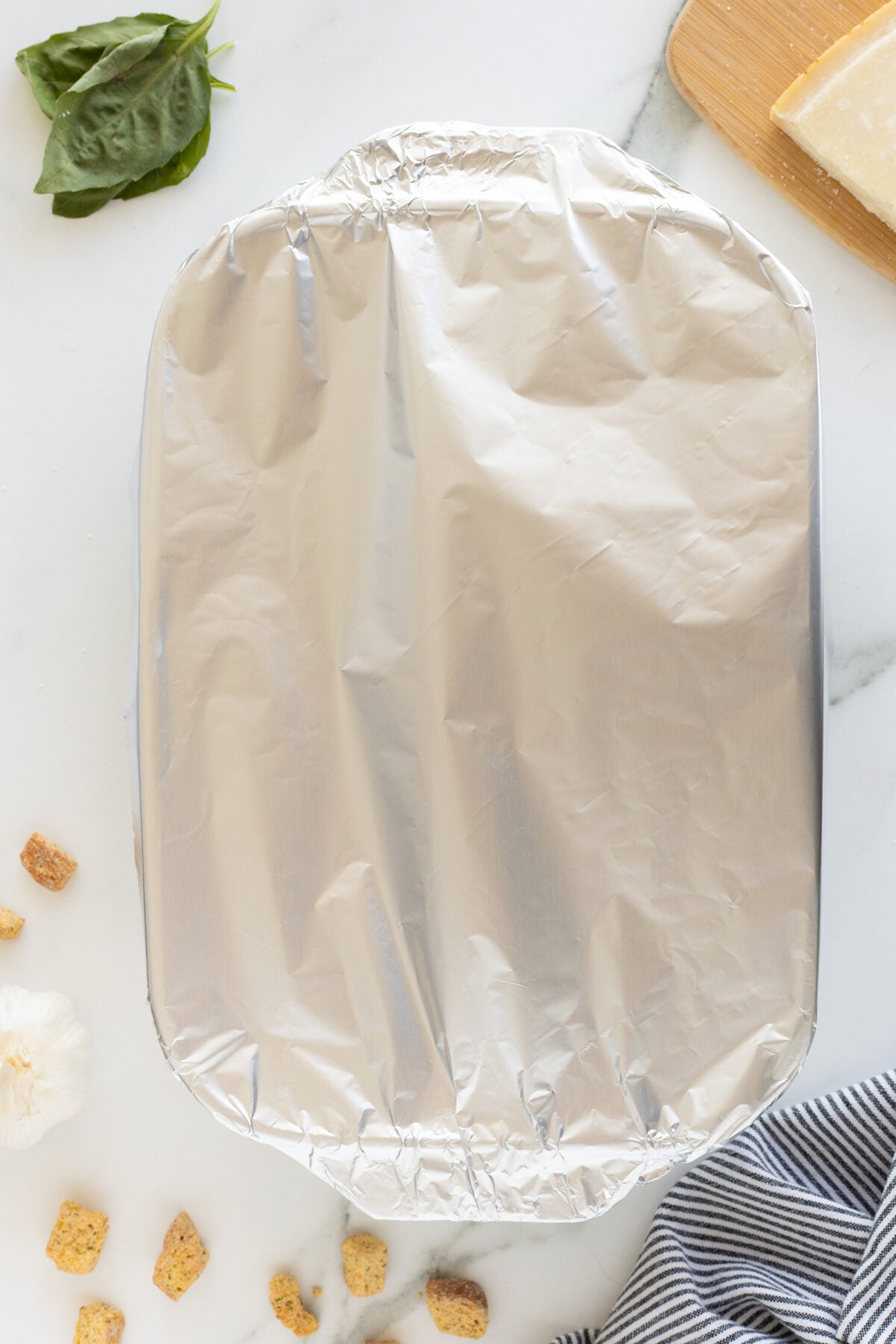 A casserole dish covered with aluminum foil sits on a white surface, surrounded by basil leaves, a block of cheese on a wooden board, croutons, a garlic bulb, and a striped cloth.