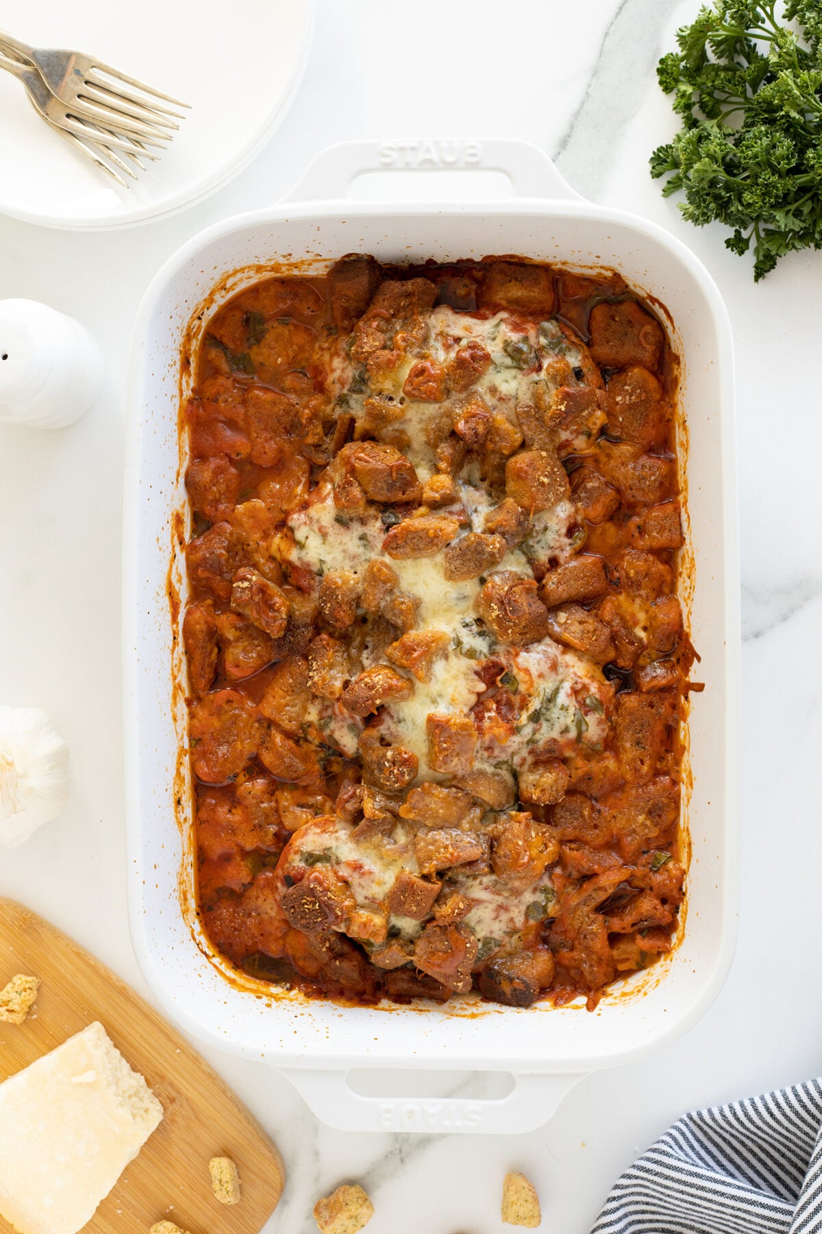 A white baking dish filled with baked Italian sausage, tomato sauce, and melted cheese sits on a marble surface, surrounded by parsley, a fork on a plate, Parmesan cheese, garlic, and croutons.