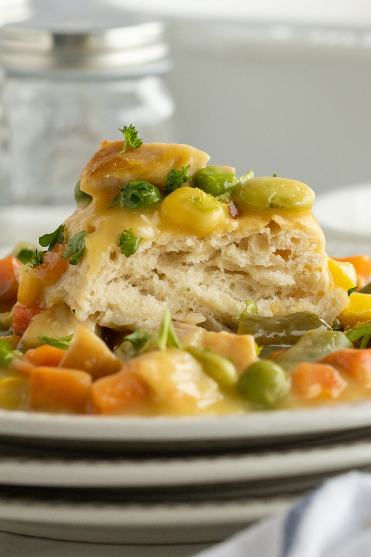 A flaky biscuit topped with creamy vegetable gravy, including carrots, lima beans, and peas, garnished with fresh herbs and served on a white plate.