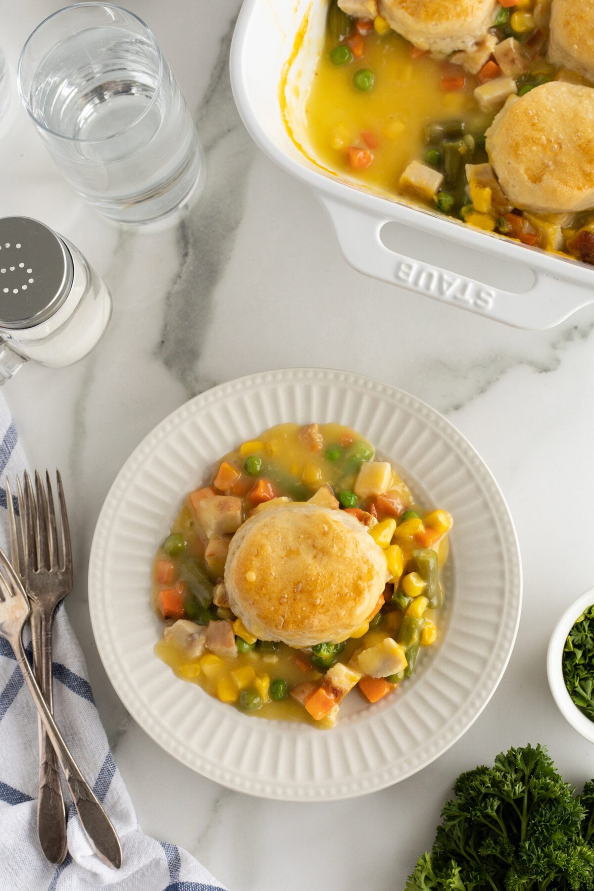 A white plate holds a biscuit atop a mixture of diced chicken, carrots, peas, and corn in a creamy sauce. Nearby are a fork and knife, a cloth napkin, and a casserole dish filled with the same meal.