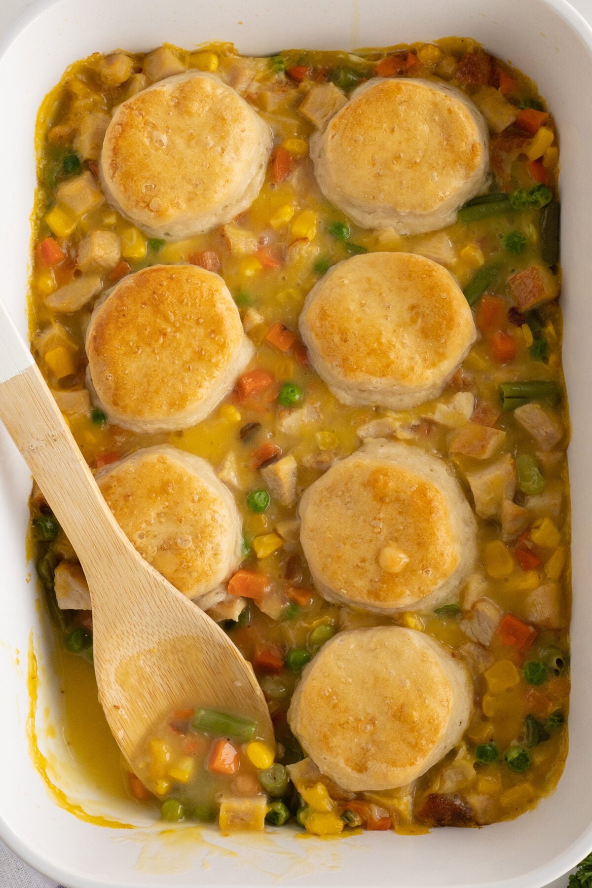 A casserole dish filled with creamy mixed vegetables and diced meat, topped with eight golden-brown biscuits, with a wooden spoon resting inside the dish.