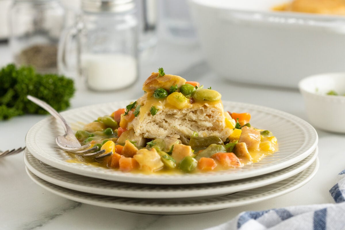 A plate of creamy vegetable pot pie filling topped with a flaky biscuit, garnished with parsley. The dish sits on stacked white plates, with a fork and kitchen items in the background.