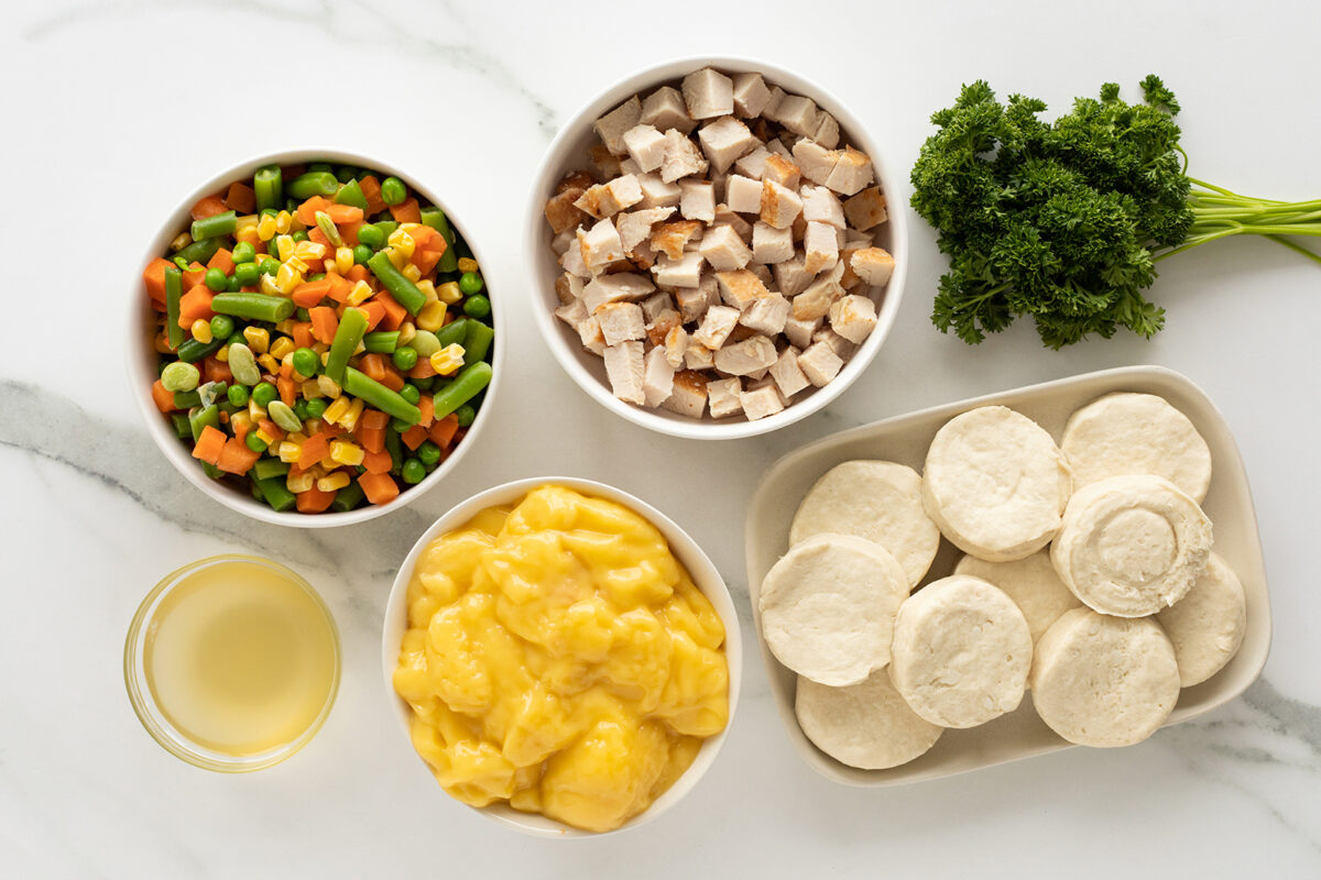 Five ingredients on a white surface: a bowl of mixed vegetables, a bowl of diced cooked chicken, a bowl of yellow sauce, a plate with raw biscuit dough, and a bunch of fresh parsley.