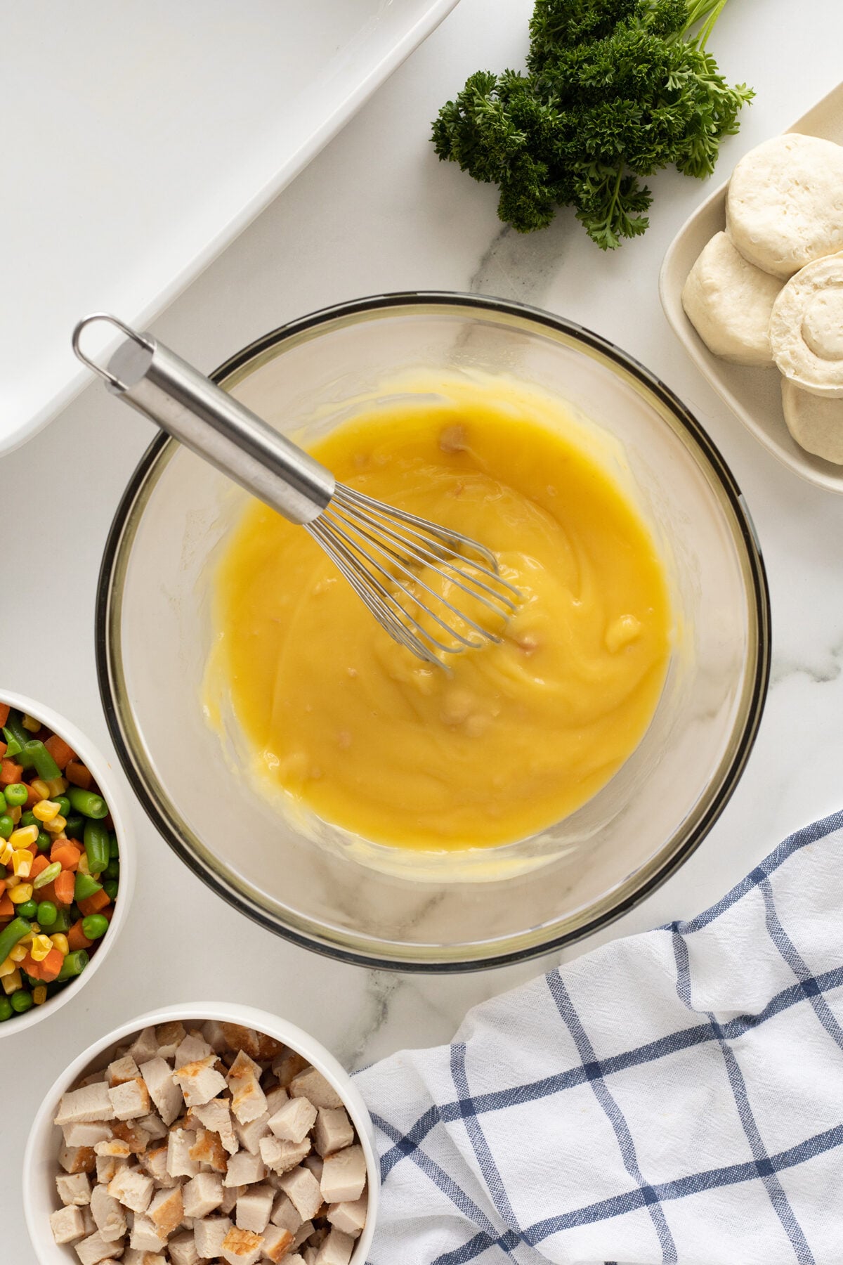 A glass bowl with yellow mixture and whisk sits on a countertop, surrounded by a plate of biscuit dough, chopped parsley, mixed vegetables, diced chicken, and a blue-and-white kitchen towel.