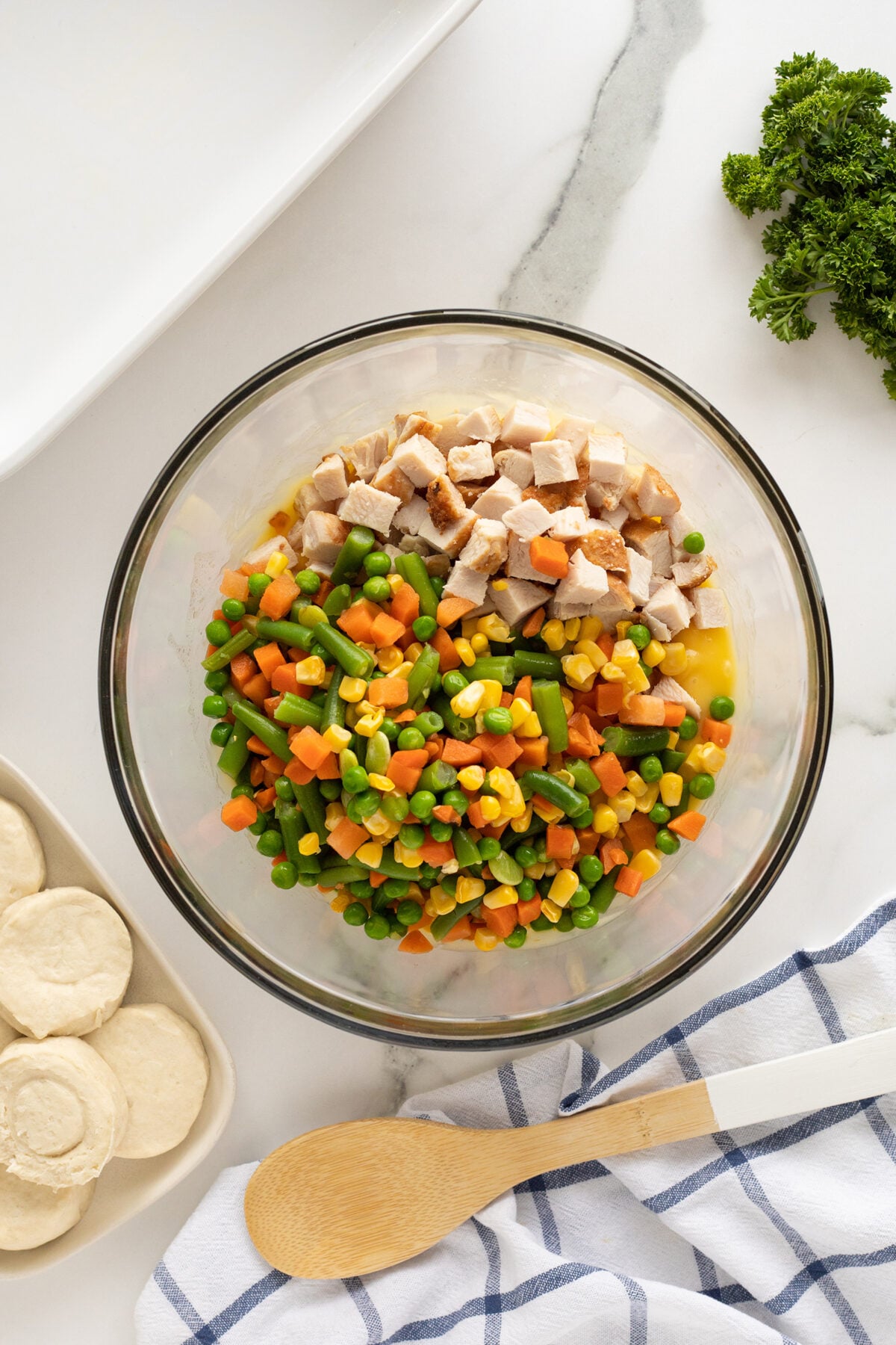 A glass bowl filled with diced chicken, mixed vegetables, and corn sits on a white marble surface. Nearby are biscuit dough rounds, a wooden spoon, a blue-striped towel, and fresh parsley.
