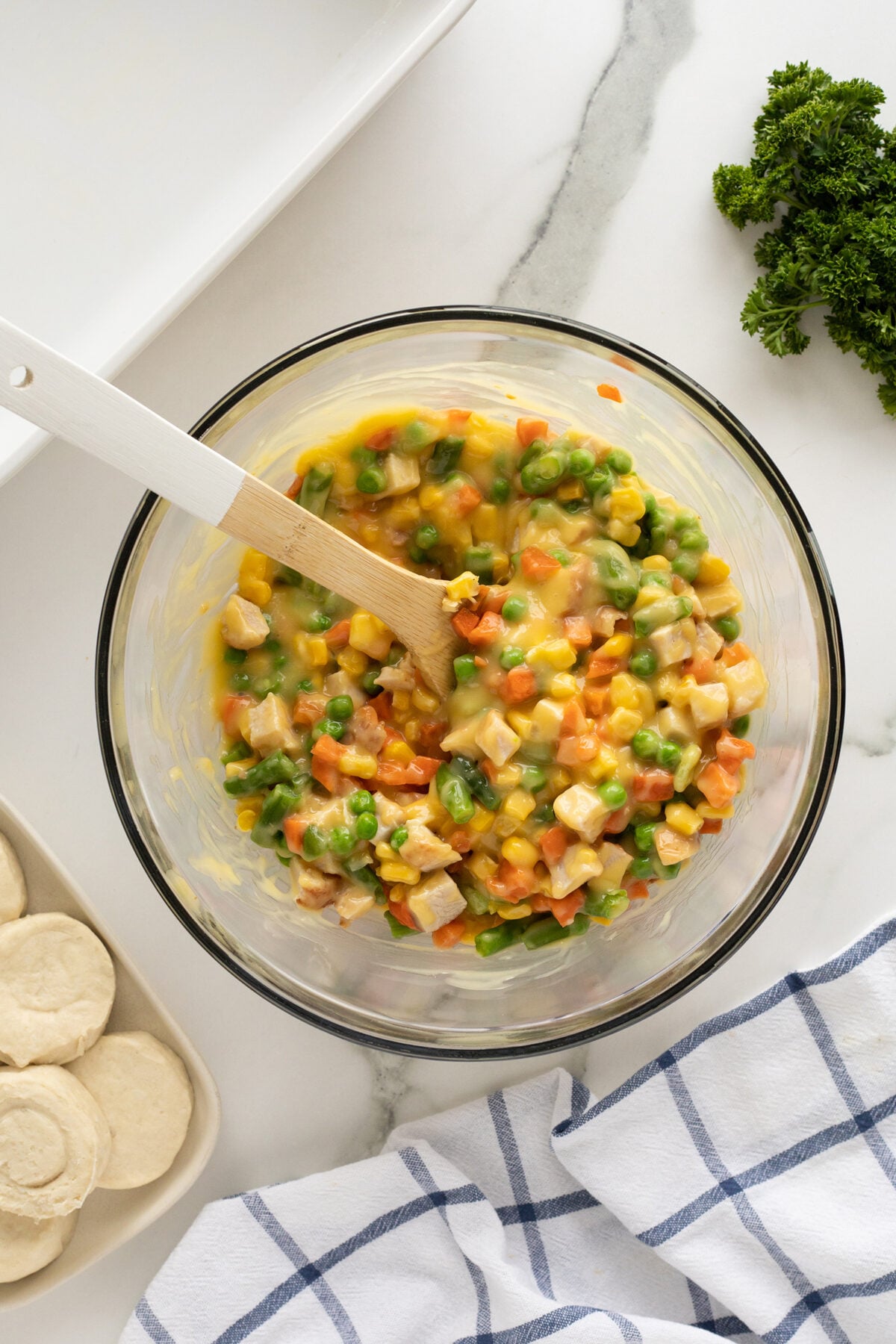 A glass bowl filled with a colorful mixture of diced vegetables and sauce, being stirred with a wooden spoon. Biscuits, parsley, and a blue-striped towel are nearby on a white countertop.