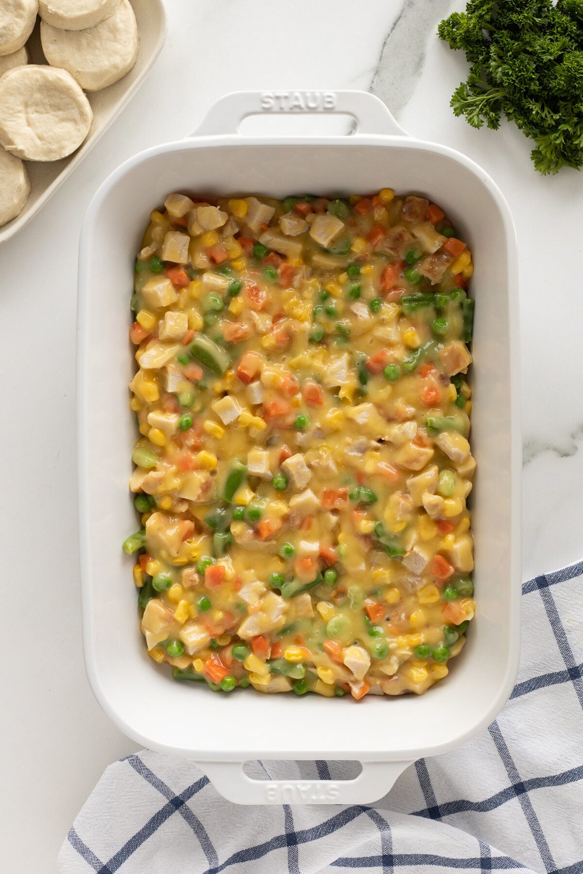 A white baking dish filled with a mixture of diced chicken, corn, carrots, peas, and green beans in a creamy sauce. Unbaked biscuits are on a plate nearby, with fresh parsley and a blue-striped towel beside them.