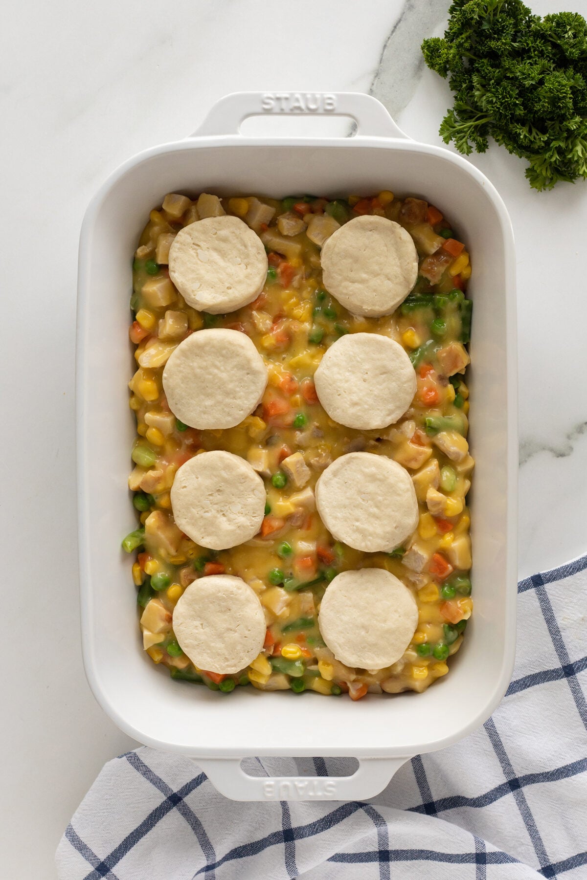 A white baking dish filled with a mixture of chicken, corn, peas, and carrots in a creamy sauce, topped with seven raw biscuit rounds. Fresh parsley and a striped towel are nearby on a marble surface.