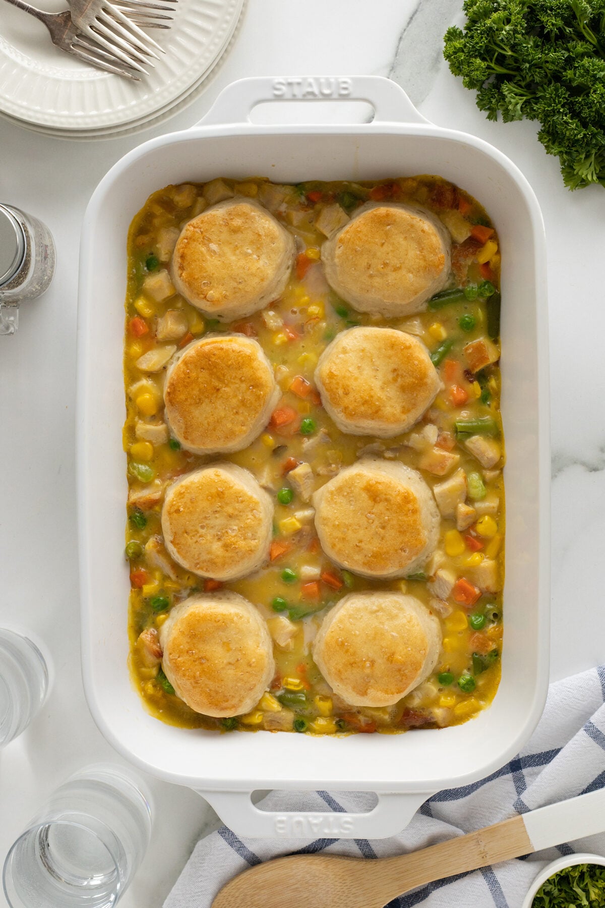 A white baking dish filled with creamy chicken and vegetable casserole, topped with eight golden-brown biscuits. Surrounding the dish are plates, utensils, parsley, and glasses on a white marble surface.