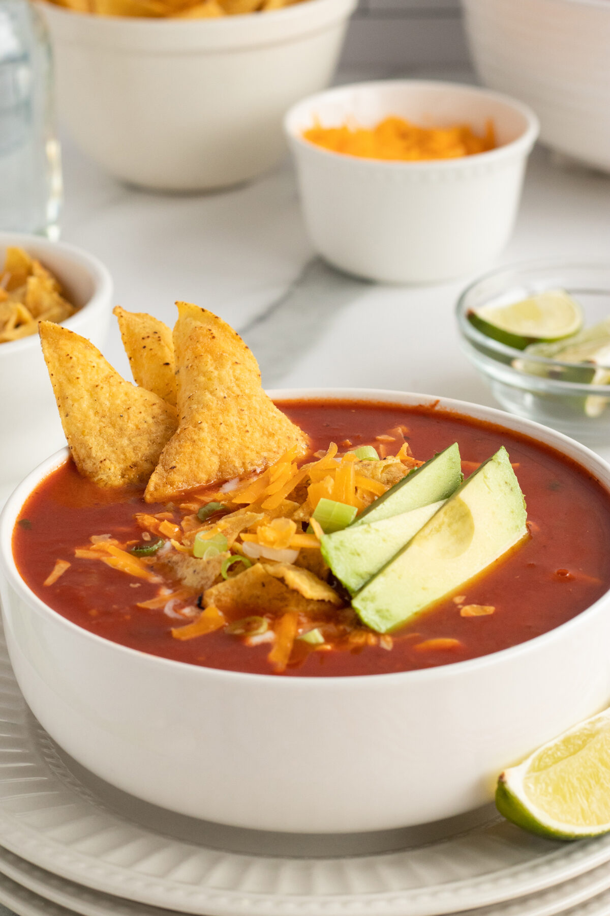 A bowl of tomato-based soup topped with shredded cheese, avocado slices, and tortilla chips, with a lime wedge on the side. Bowls of chips, cheese, and lime wedges are in the background.