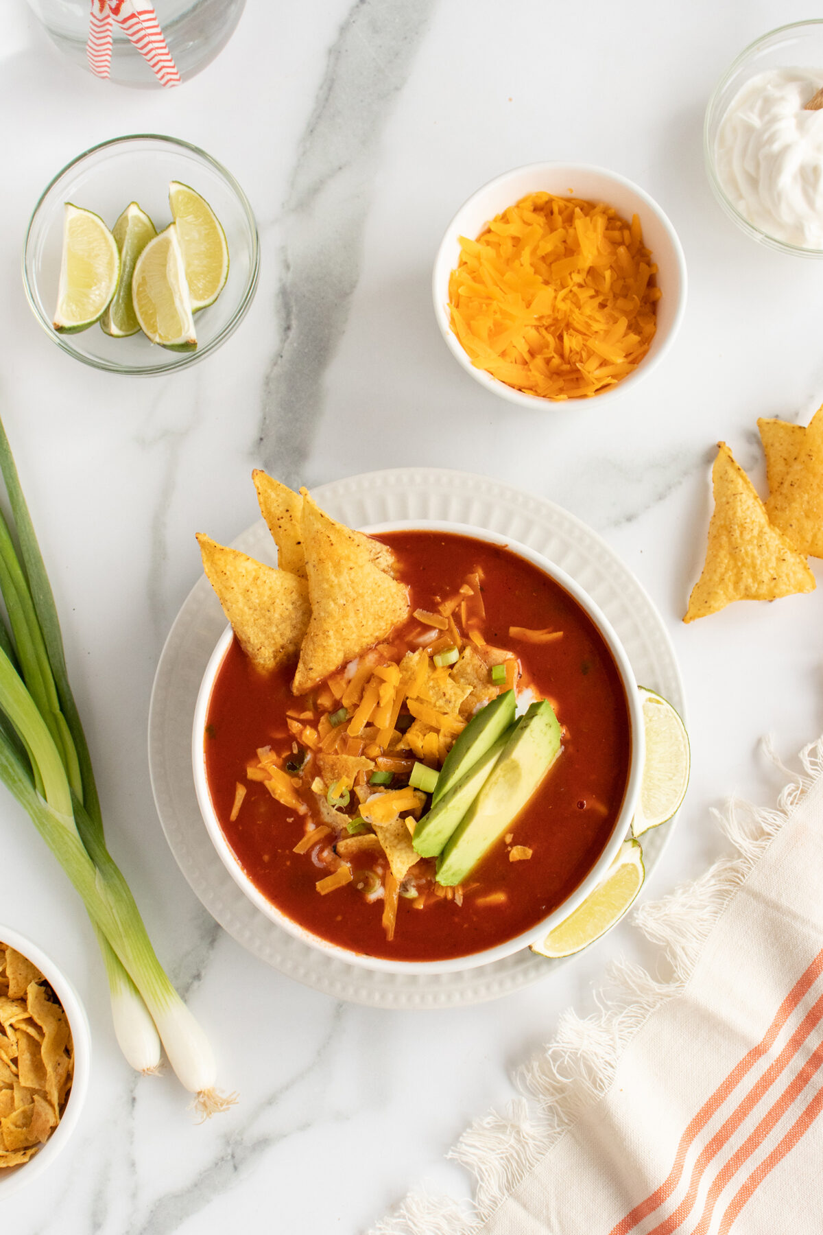 A bowl of tortilla soup topped with avocado slices, shredded cheese, and tortilla chips, surrounded by lime wedges, green onions, sour cream, extra cheese, and a striped napkin on a marble surface.