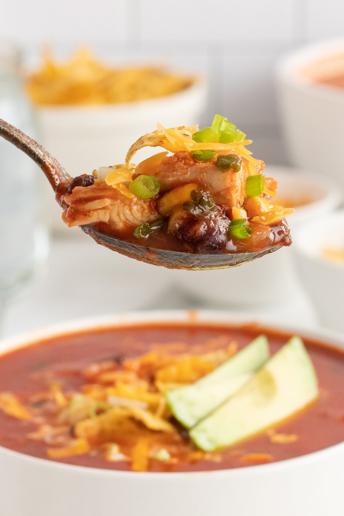 A close-up of a spoonful of chicken tortilla soup topped with shredded cheese and green onions, held above a bowl filled with soup and garnished with sliced avocado.