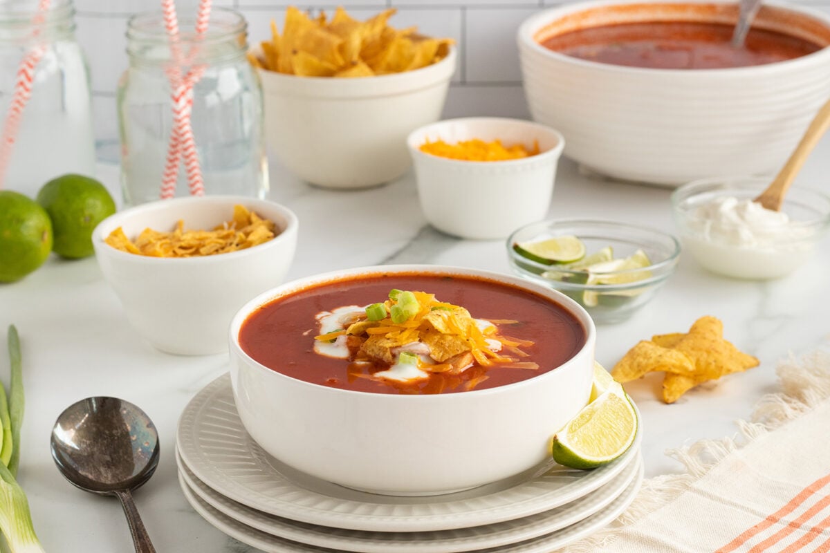 A bowl of tomato-based soup topped with tortilla chips, cheese, and green onions sits on a plate with a lime wedge. Surrounding it are bowls of chips, cheese, sour cream, lime slices, and glasses of water with straws.