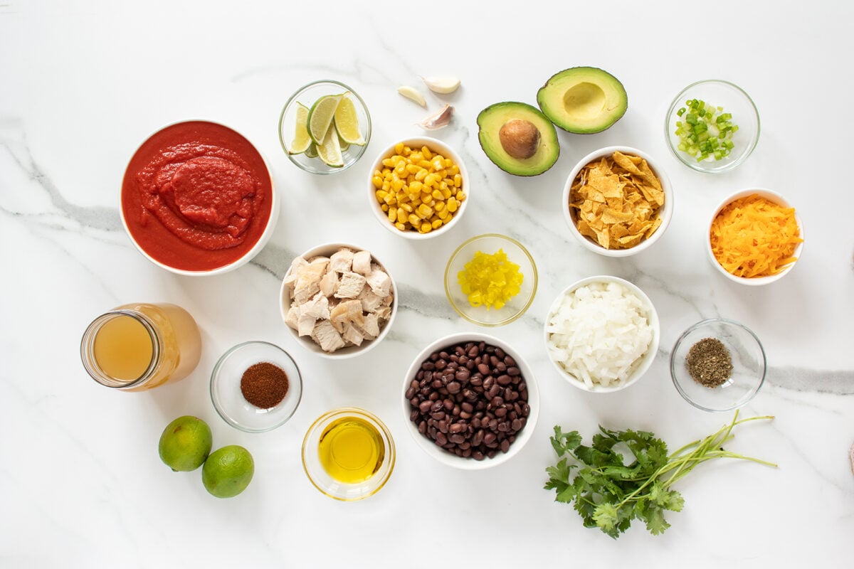 Assorted ingredients for a Mexican-inspired dish arranged on a white marble surface, including tomato sauce, corn, avocado, tortilla chips, cheese, black beans, chicken, onions, limes, cilantro, and various seasonings.