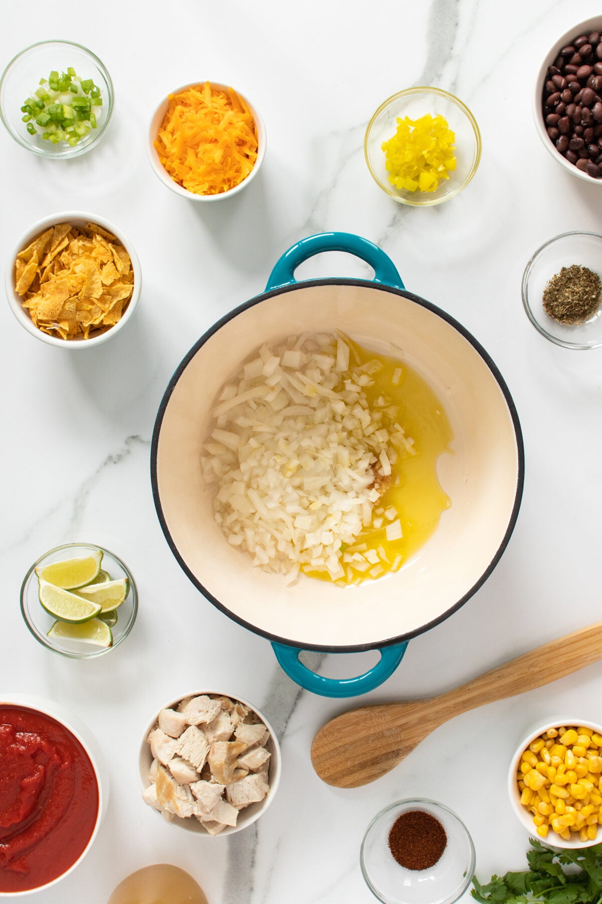 A pot with chopped onions and melted butter sits on a marble surface, surrounded by bowls of various ingredients including beans, corn, chicken, cheese, lime wedges, chips, peppers, spices, and tomato sauce.
