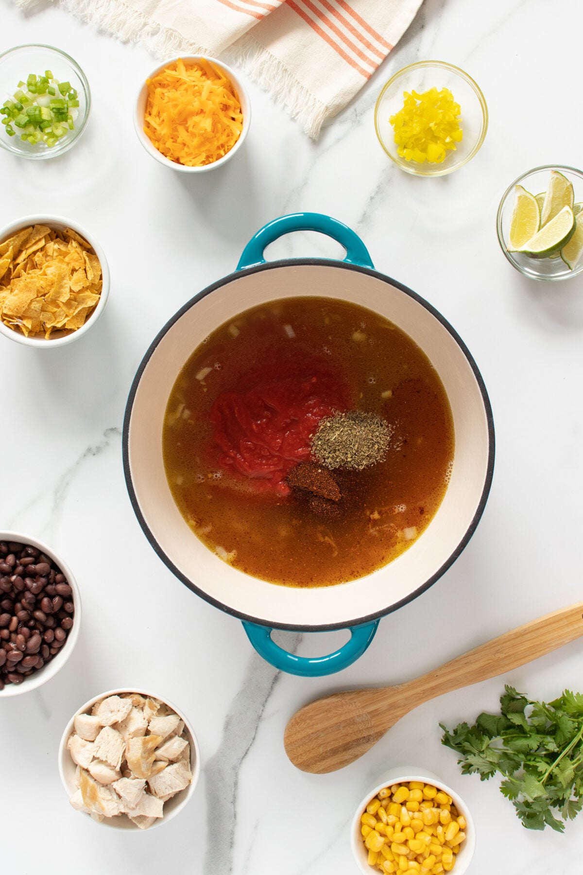 A pot with broth, tomatoes, and seasonings sits on a marble counter surrounded by bowls of black beans, chicken, corn, tortilla chips, cheese, scallions, jalapeños, lime wedges, and fresh cilantro. A wooden spoon is nearby.