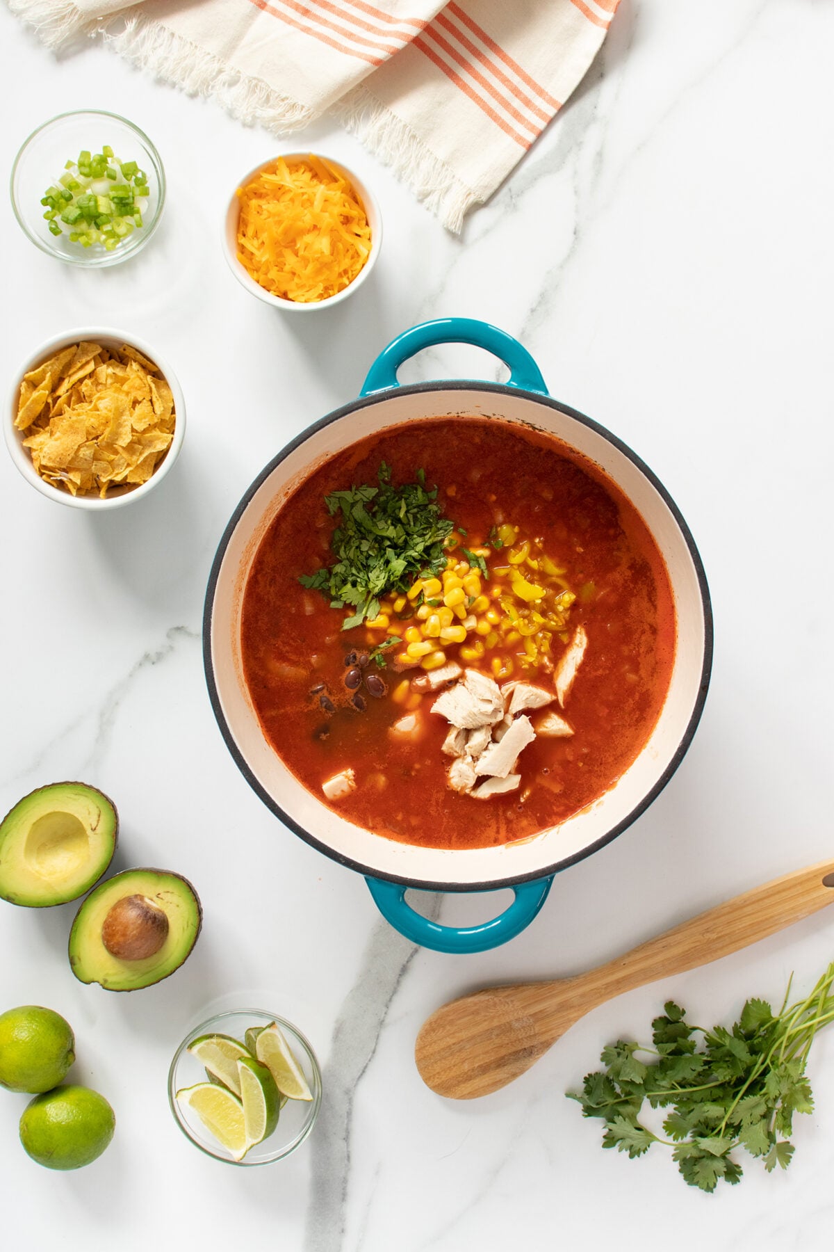 A pot of tortilla soup with chicken, corn, and cilantro sits on a marble counter. Surrounding it are small bowls of cheese, tortilla strips, green onions, avocado halves, limes, and fresh cilantro.