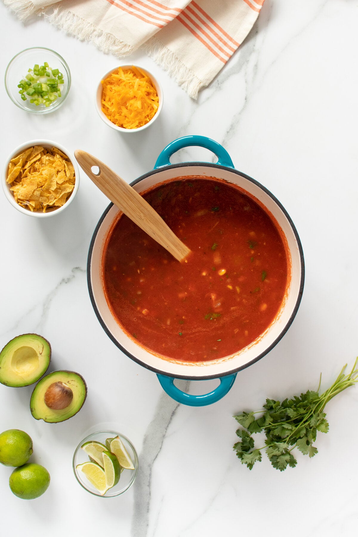 Overhead view of a pot of tomato-based soup with a wooden spoon, surrounded by bowls of shredded cheese, chopped green onions, tortilla chips, halved avocado, limes, cilantro, and a striped towel on a marble surface.