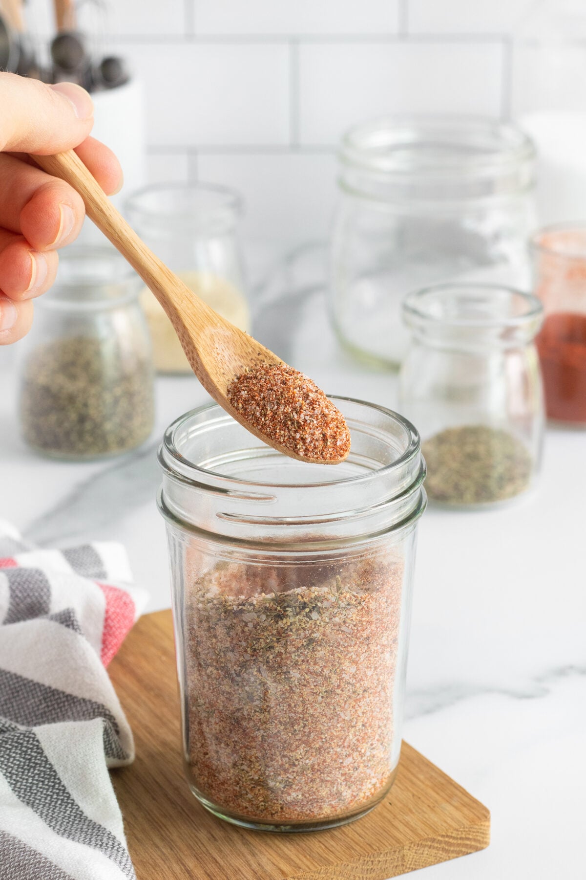 A hand holds a wooden spoon with a scoop of seasoning mix above a glass jar filled with the same mix. Other jars with spices and a striped cloth are in the background on a white countertop.