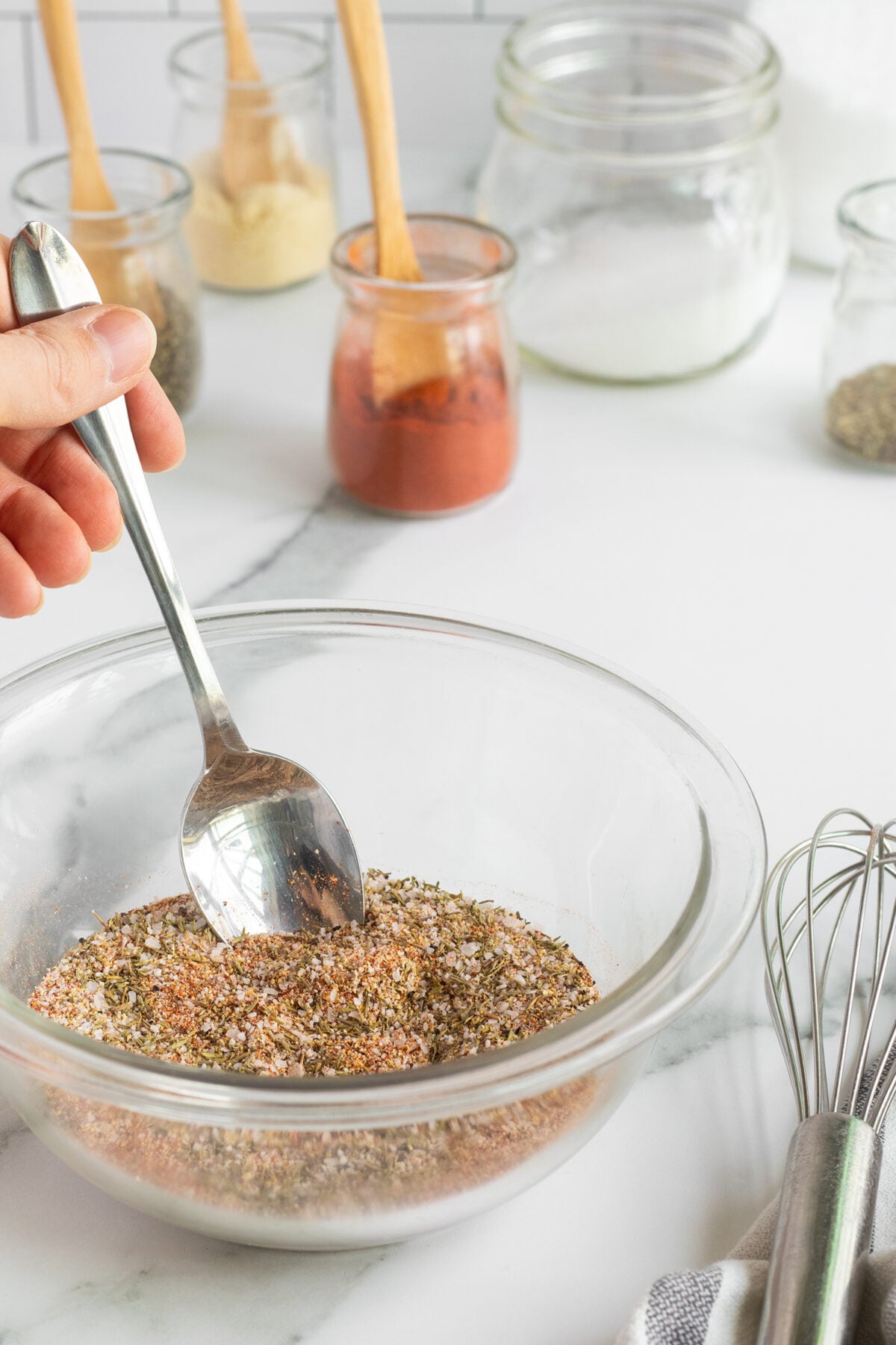 A hand holds a spoon stirring a blend of spices in a glass bowl on a white countertop, with jars of ingredients and a whisk nearby.