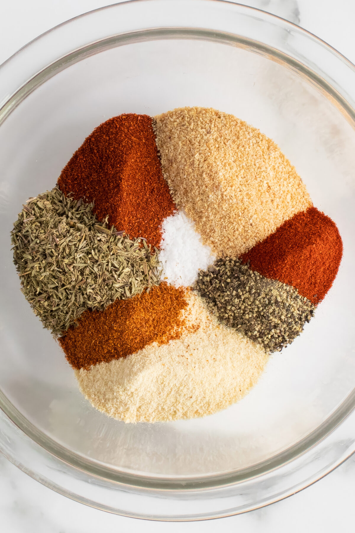 A clear glass bowl filled with separate piles of various ground spices and herbs, including paprika, black pepper, salt, thyme, and garlic powder, arranged in a circular pattern on a white surface.