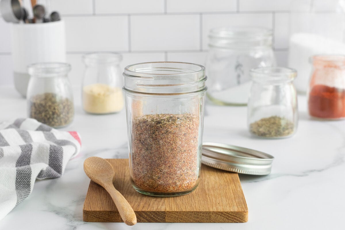 A glass jar filled with a mixed seasoning blend sits on a wooden board with a wooden spoon beside it. The jar lid is off, and various spice jars are blurred in the background on a white countertop.