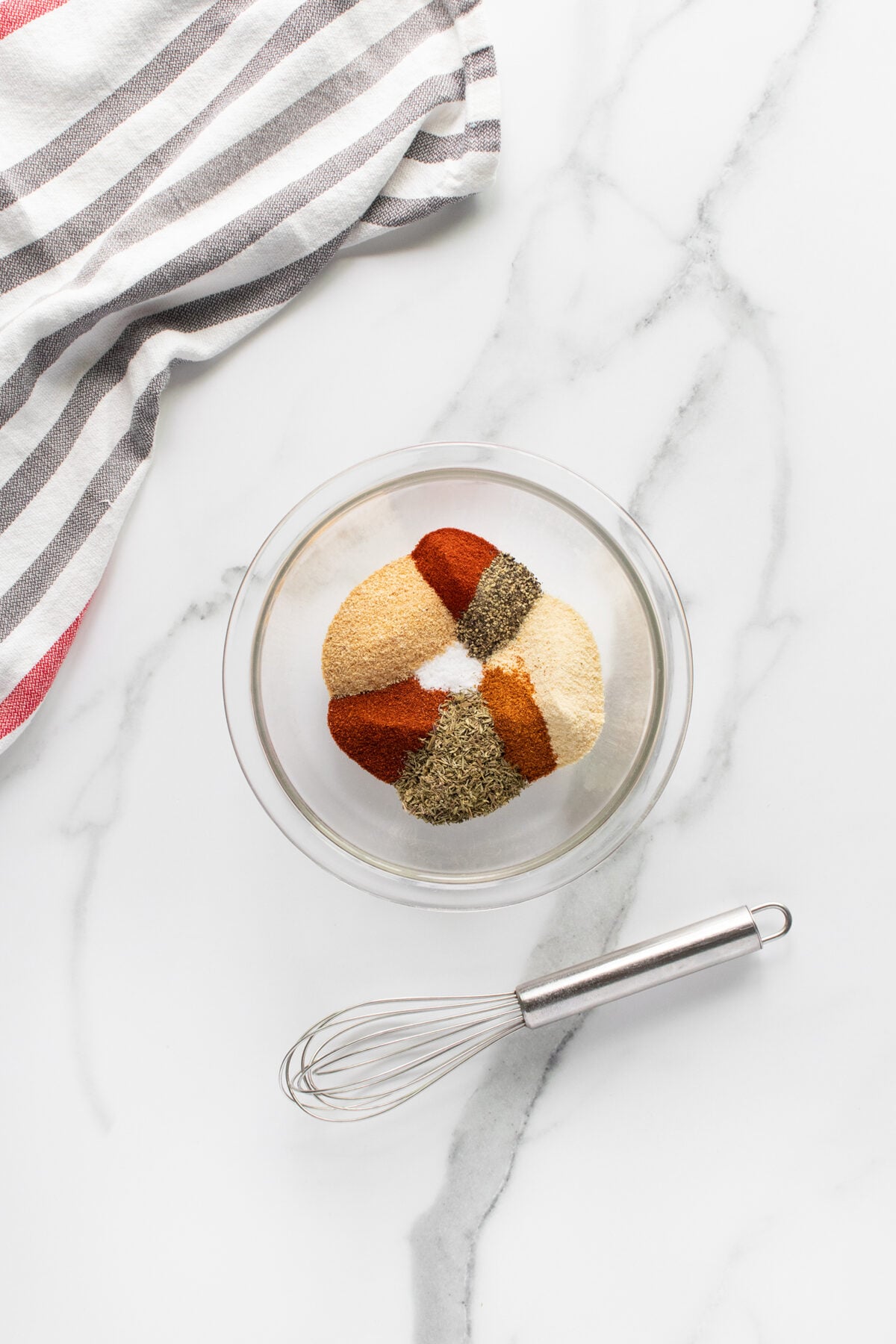 A glass bowl with assorted spices arranged in sections, next to a metal whisk on a white marble surface. A striped kitchen towel is partially visible in the top left corner.