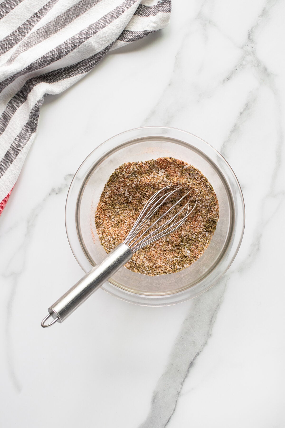 A glass bowl filled with a mix of dry spices and a metal whisk rests on a white marble surface, with a striped kitchen towel partially visible in the corner.