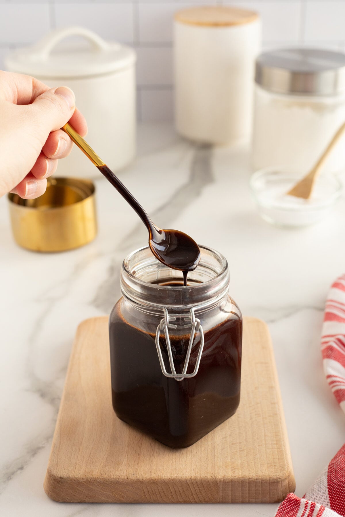 A hand holds a spoon dripping with dark syrup above a glass jar filled with the same syrup, placed on a wooden board on a white countertop. Kitchen containers and utensils are in the background.
