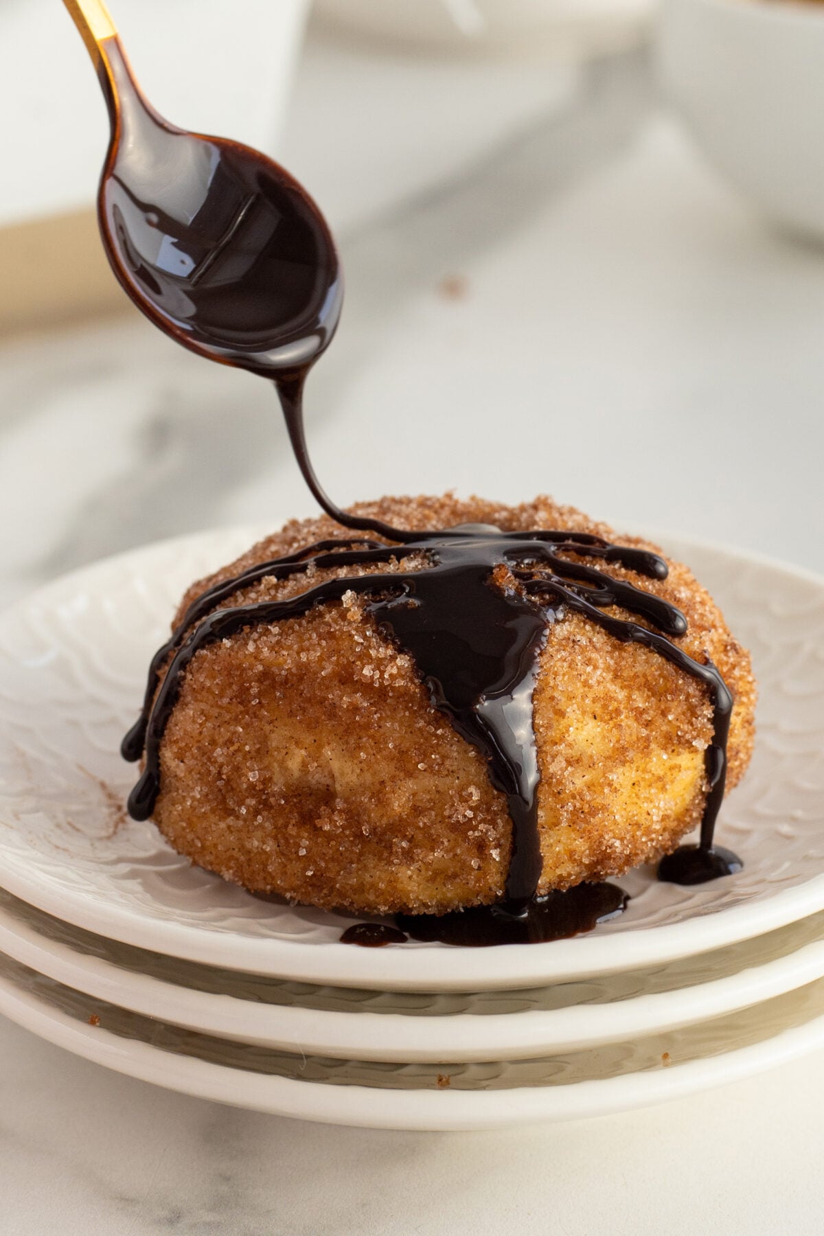 A hand drizzles chocolate sauce from a spoon over a round, cinnamon sugar-coated pastry on a stack of white plates.