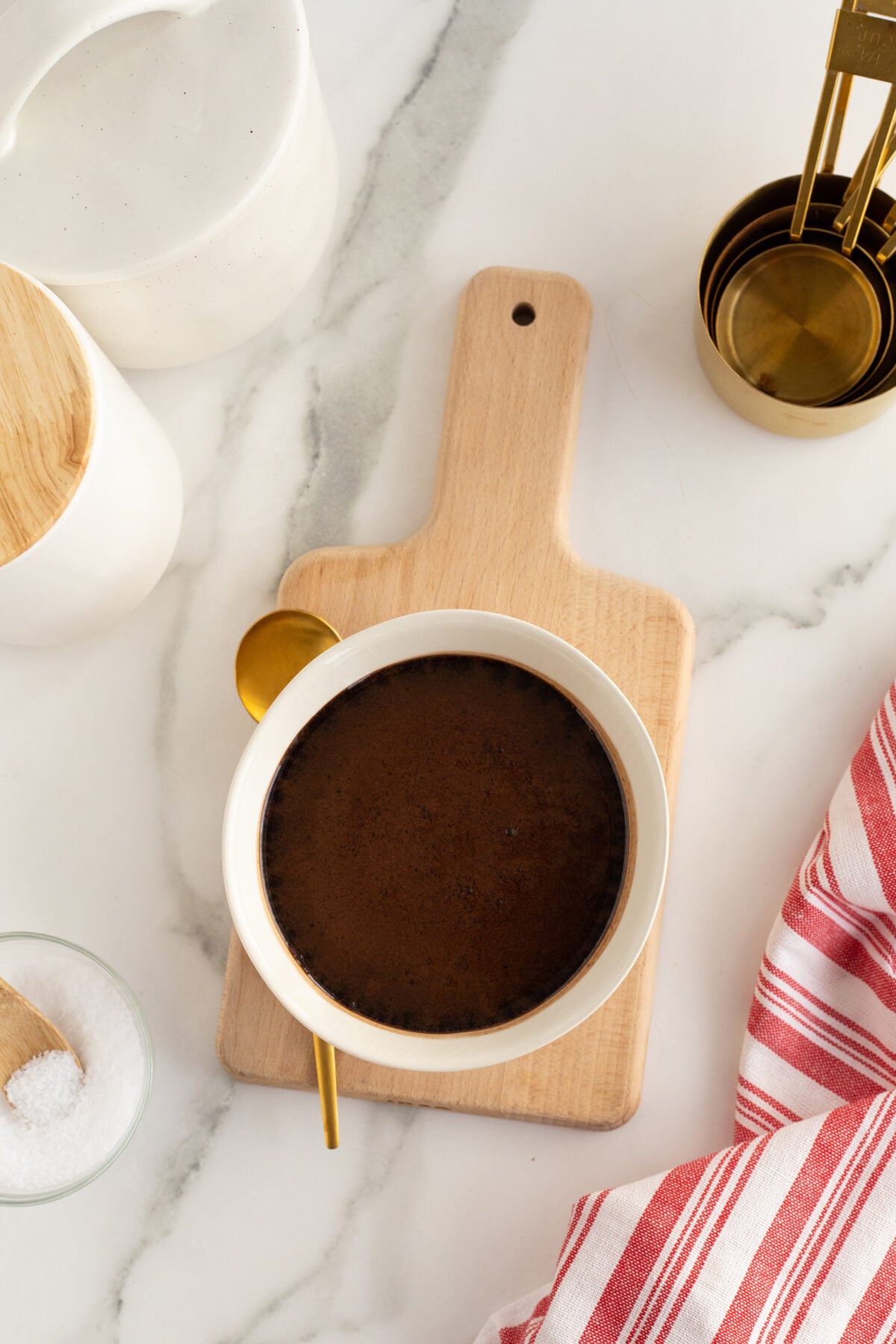 A bowl of chocolate mixture with a gold spoon rests on a wooden cutting board. Nearby are a jar with a wooden lid, a small bowl of salt, gold measuring cups, and a red-striped cloth on a white marble surface.