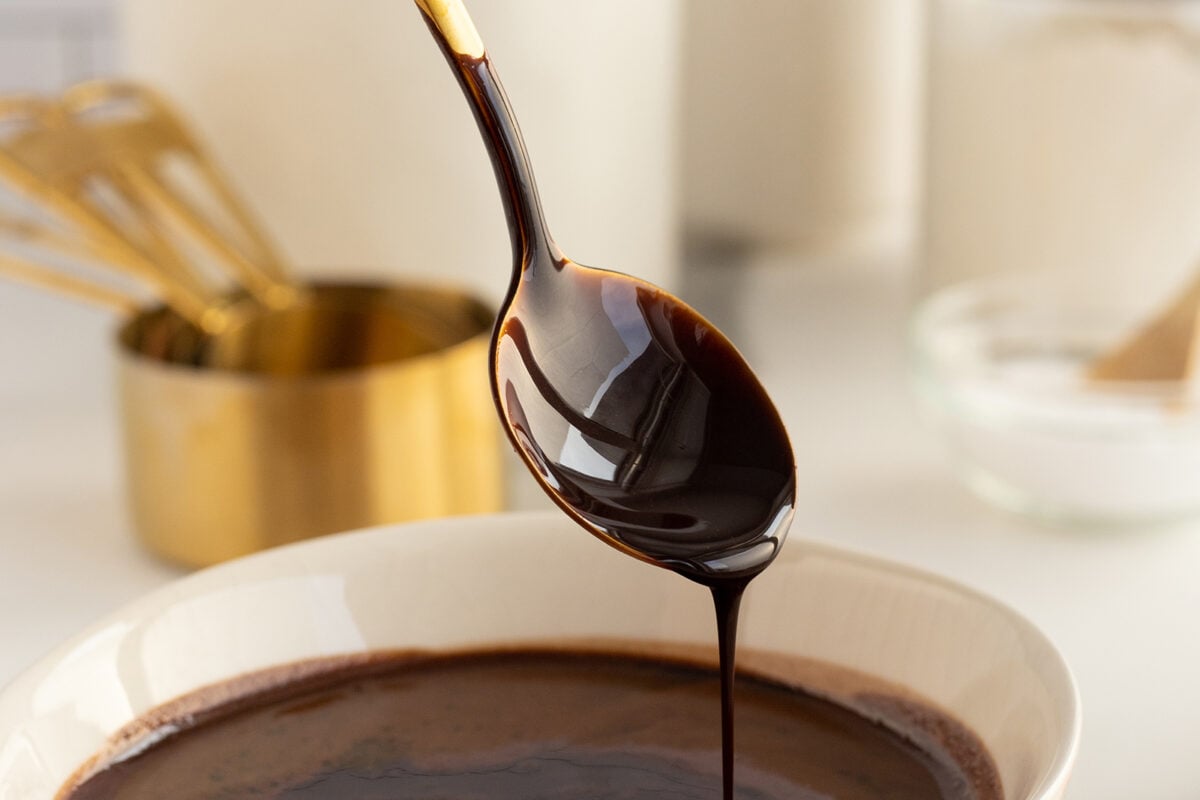 A spoon drizzles thick, glossy chocolate sauce over a white bowl filled with more sauce. In the blurred background, gold measuring cups and a glass bowl are visible on a white surface.