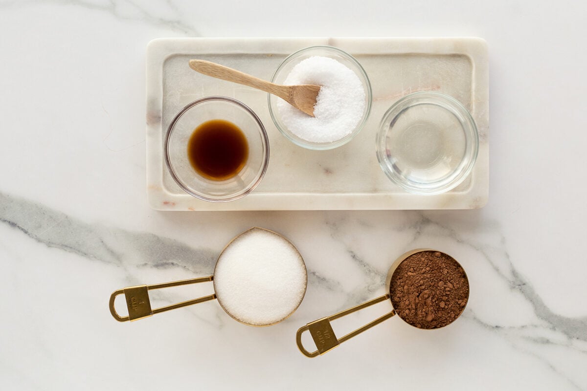 An overhead view of baking ingredients: a bowl of salt with a wooden spoon, a bowl of clear liquid, a bowl of brown liquid, a measuring cup of sugar, and a measuring cup of cocoa powder on a marble surface.