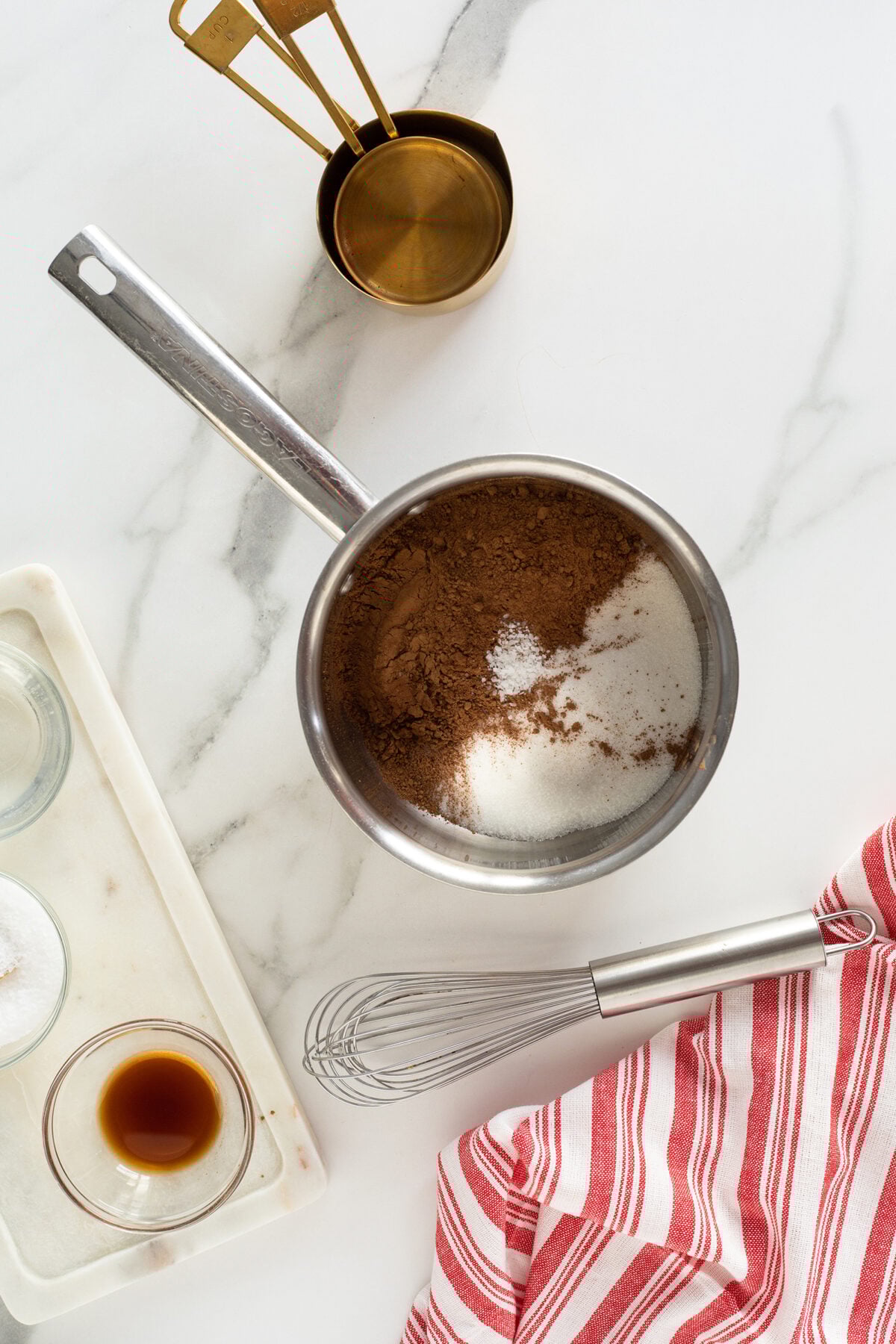 A saucepan filled with cocoa powder, sugar, and salt sits on a marble counter next to a whisk, a red-striped cloth, measuring spoons, a small bowl of vanilla, and a tray with other ingredients.