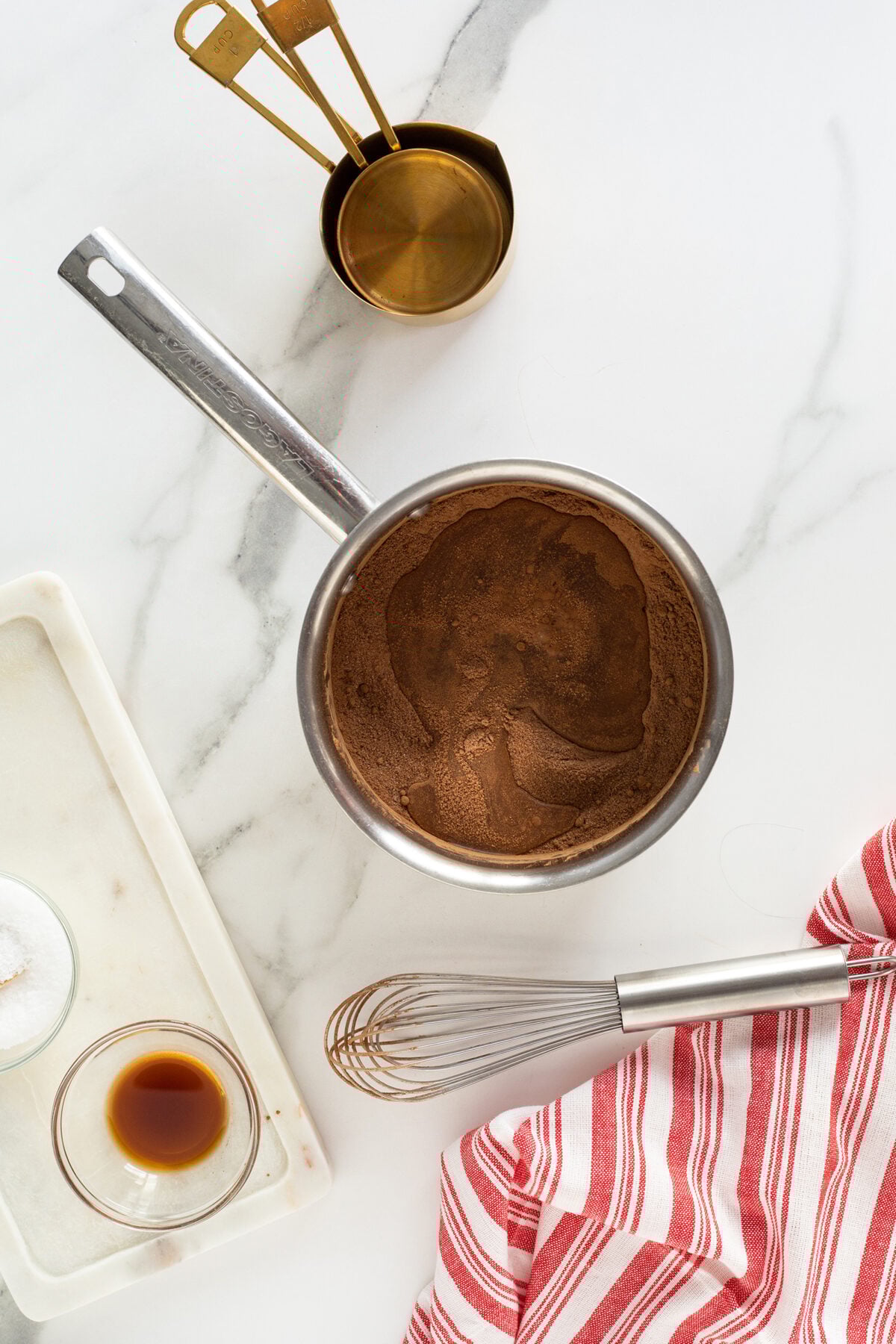A saucepan filled with chocolate mixture sits on a marble countertop beside a metal whisk, a red-striped cloth, measuring cups, a small bowl of vanilla, and a dish with salt.