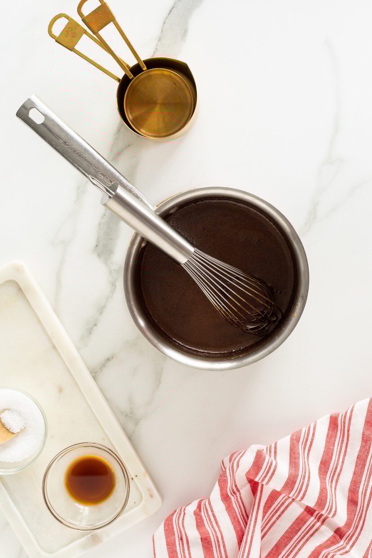 A whisk rests in a metal bowl filled with chocolate batter on a marble counter, beside brass measuring cups, a red-striped towel, a small bowl of vanilla, and a dish of sugar.
