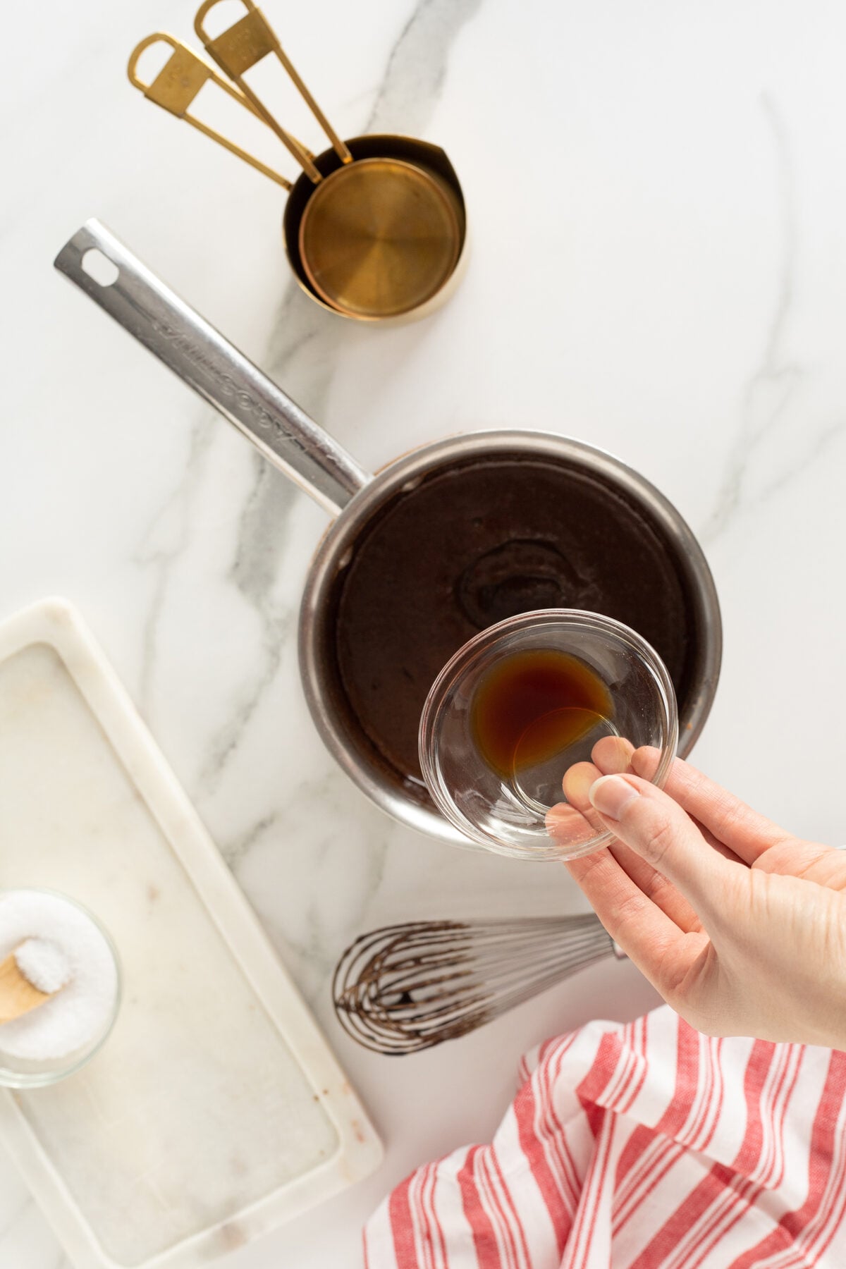 A hand pours vanilla extract from a small glass bowl into a saucepan of chocolate mixture on a white marble surface, with measuring cups, a whisk, and a red-striped towel nearby.
